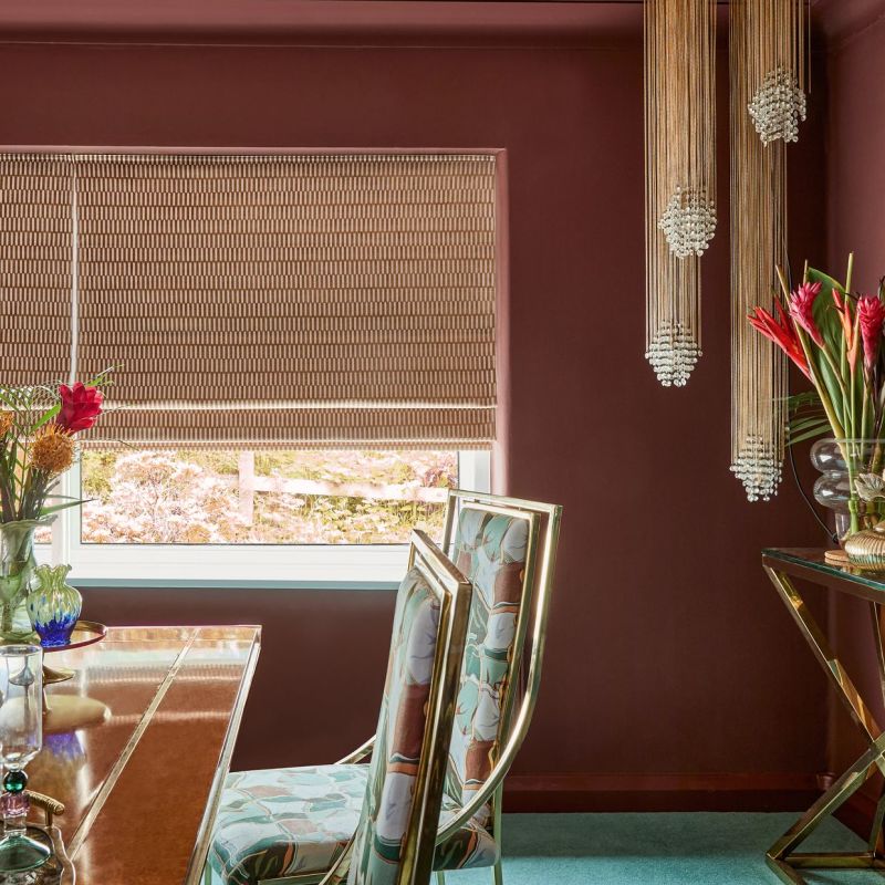 Woven beige patterned roman blind lowered over a window, filtering daylight with its bottom slightly raised; in a burgundy dining room with brass-framed floral chairs, wooden table, and beaded pendant lights.