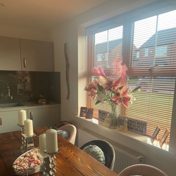Wooden horizontal blinds over a window, slats tilted to filter warm sunlight; vase of pink lilies on the sill, wooden dining table with candles in a cozy kitchen-diner.
EAT and be MERRY