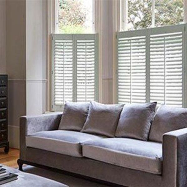 White plantation-style café shutters covering the lower halves of tall windows, slatted panels partially closed and filtering daylight; grey sofa with cushions in a lit living room, trees visible outside.
