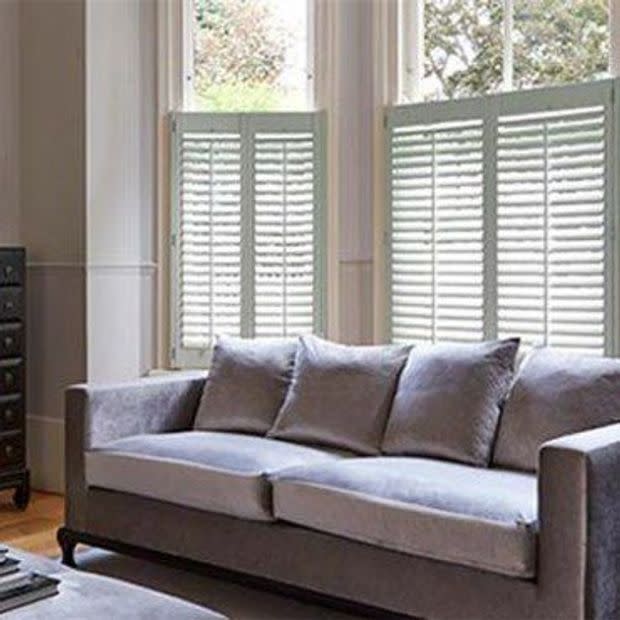 White plantation-style cafÃ© shutters covering the lower halves of tall windows, slatted panels partially closed and filtering daylight; grey sofa with cushions in a lit living room, trees visible outside.