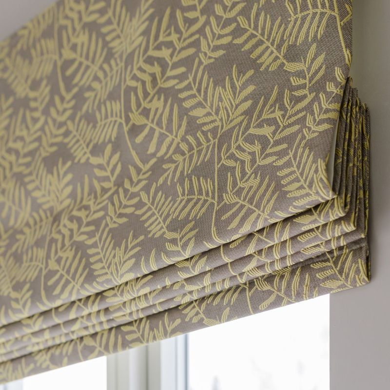 Roman blind with beige fabric and yellow fern-like pattern, neatly folded into pleats; lowered over a window, partially blocking and softly filtering daylight in a neutral-toned room.