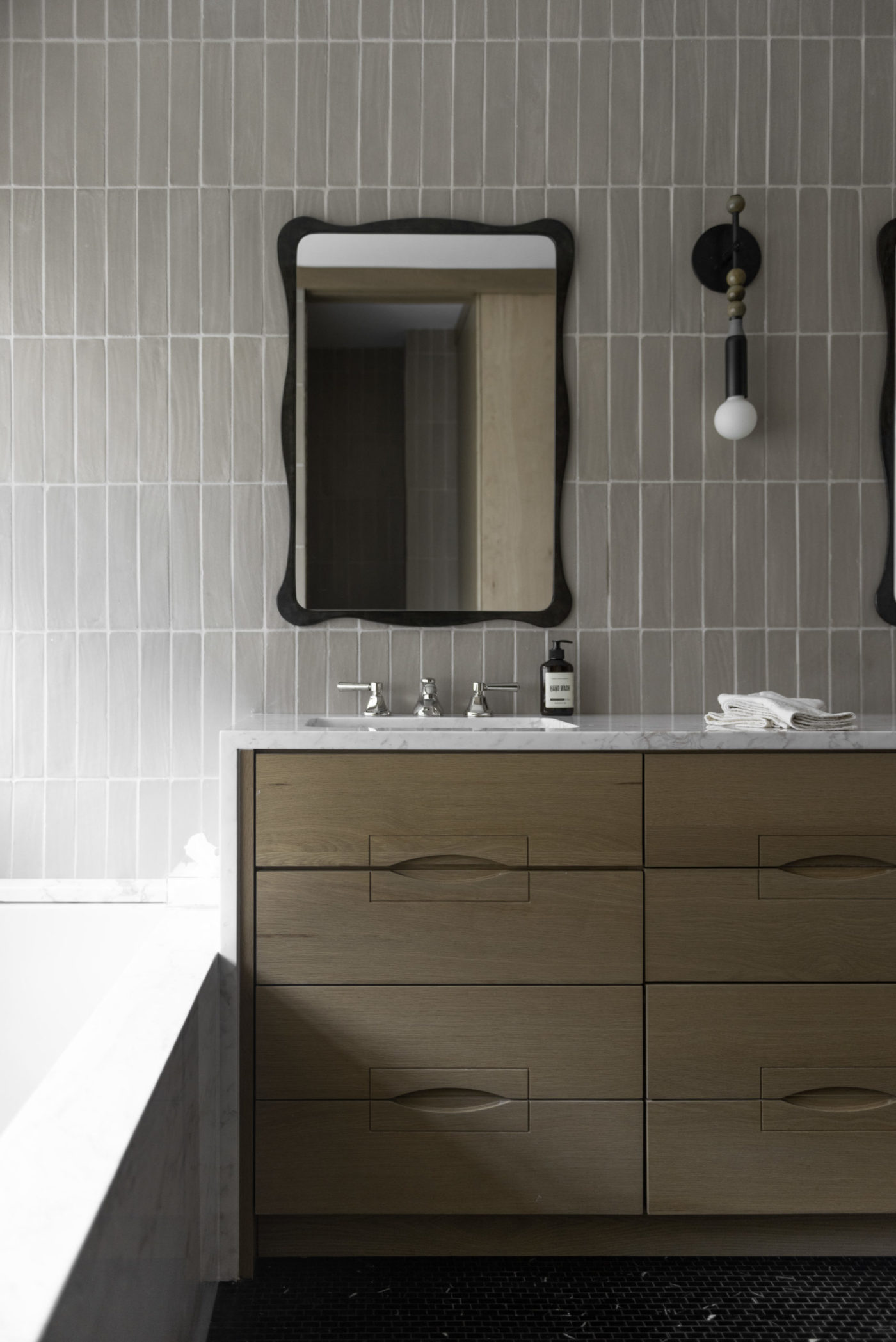 a grey tiled bathroom wall with two sinks and two mirrors.