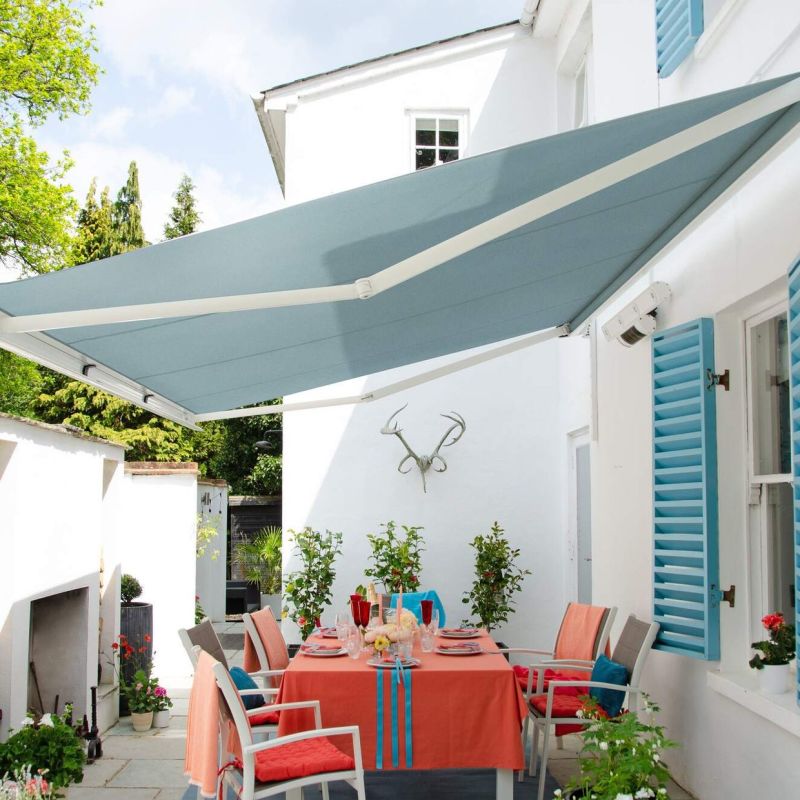 Blue fabric retractable awning mounted above a patio, fully extended with white support arms, shading a coral-tablecloth dining table beneath; sunny white-walled courtyard with turquoise shutters and potted plants.