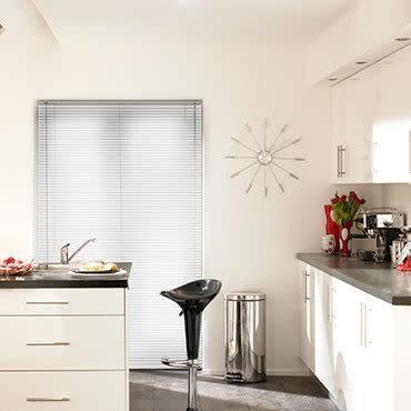 Horizontal aluminum Venetian blinds covering a window, closed and diffusing daylight; in a bright modern white kitchen with dark countertops, chrome barstool, stainless bin, sunburst wall clock and red tulips.