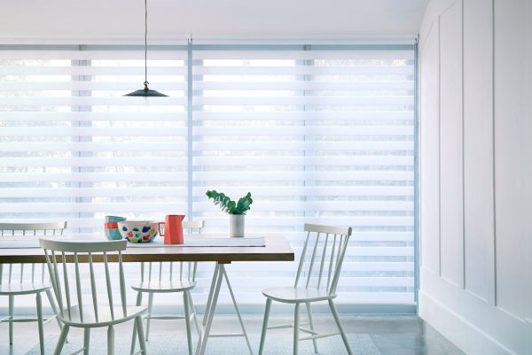 Zebra blinds cover floor-to-ceiling windows, alternating translucent and opaque stripes lowered to softly filter daylight, in a bright minimalist dining room with a wooden table, white chairs and a plant.