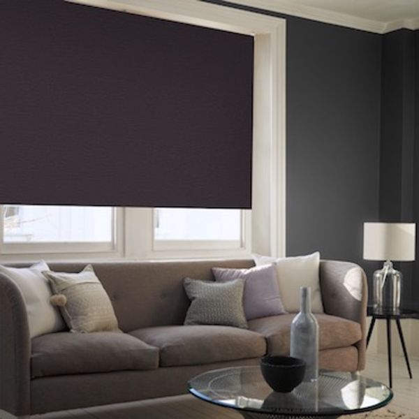Dark fabric roller blind lowered nearly to the sill, blocking light over a two-pane living-room window; charcoal walls, beige sofa with cushions, glass coffee table and side lamp.