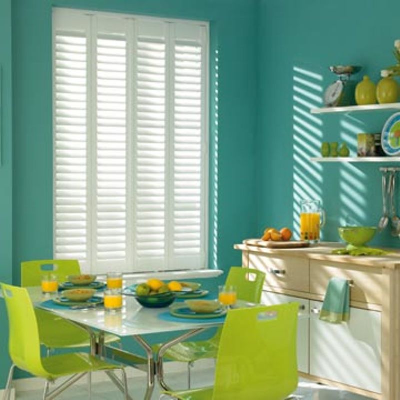 White plantation shutters covering a window, slatted panels mostly closed, filtering sunlight into striped shadows across a teal dining-kitchen with lime-green chairs, a set breakfast table, and wooden cabinets.