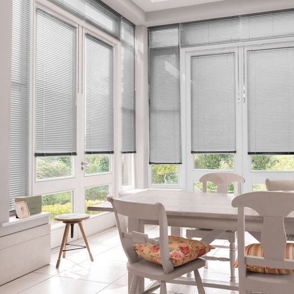 Grey horizontal Venetian blinds cover tall glass doors and windows, slats mostly closed and filtering daylight; bright white dining area with wooden table, chairs, floral cushions and garden view.