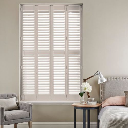 White full-height plantation shutters, four-panel with adjustable louvers, closed and filtering daylight, covering a recessed window; neutral bedroom corner with armchair, bedside table, lamp, and flowers.