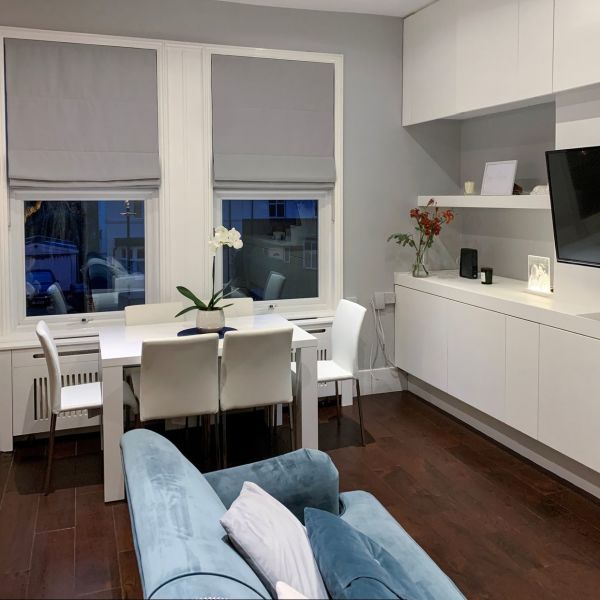 Two gray Roman fabric blinds, lowered with flat folds, partially covering windows and blocking light; modern dining-living room with white table and chairs, orchid, blue sofa, built-in cabinets, dusk.