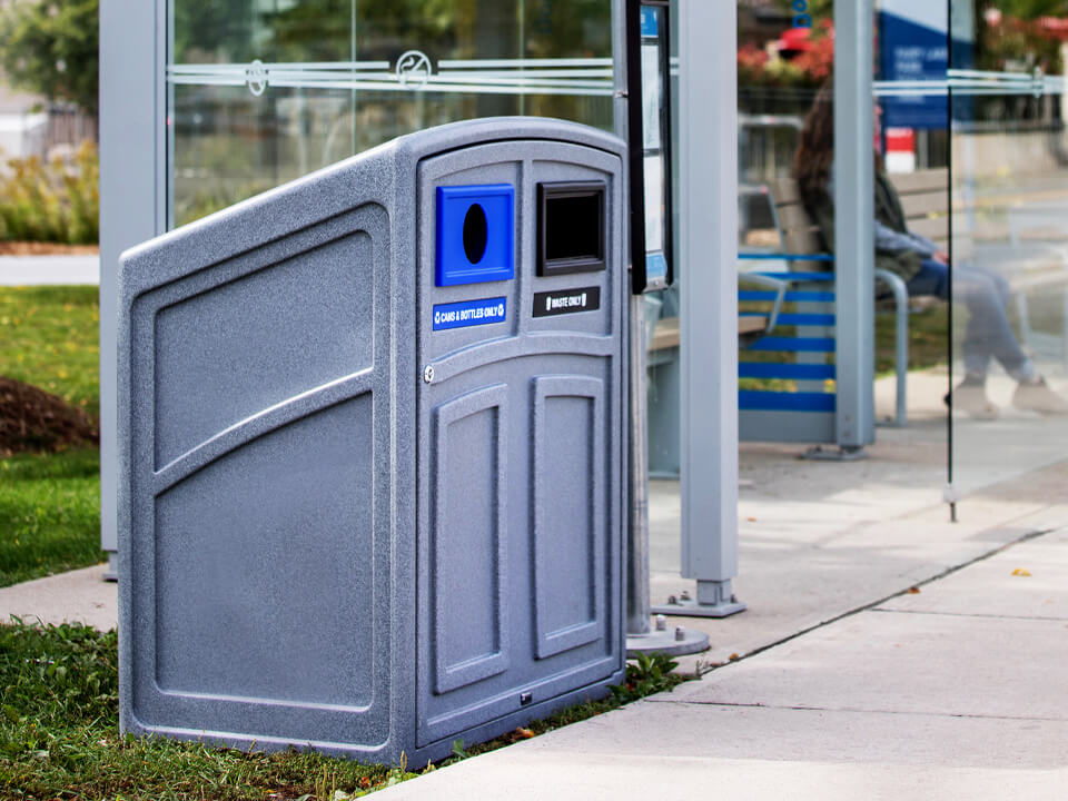 greystone plastic bin for collecting trash and recycling sitting outdoors beside city bus shelter