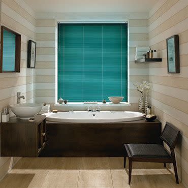 Teal horizontal blinds covering a centered window, fully lowered and filtering daylight above a built-in wooden bathtub in a striped-tile bathroom with mirrors, shelves, vase, and a chair.