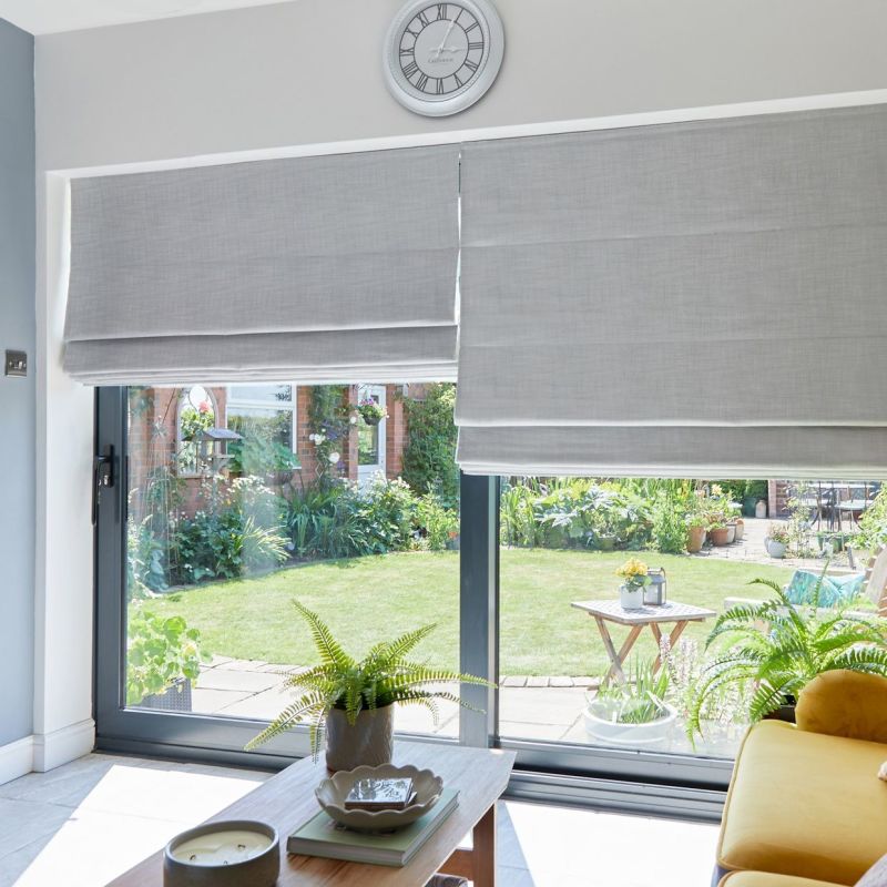 Grey Roman blinds covering sliding glass doors, lowered and overlapping slightly, filtering daylight; a coffee table with potted ferns and a yellow sofa face a sunlit lawn and flowered garden outside.

Text: Greenwich