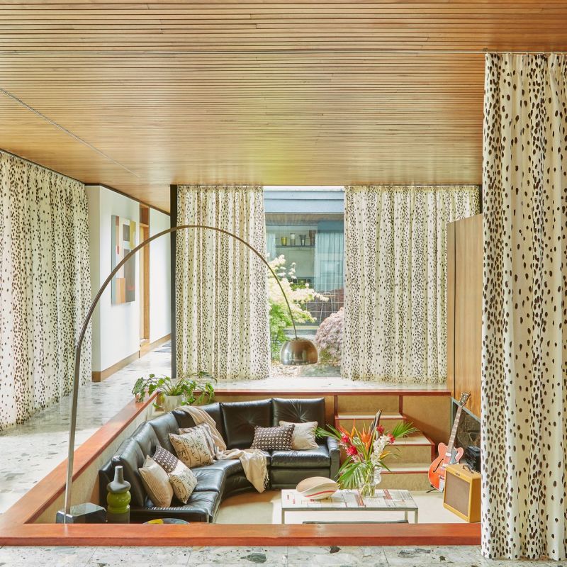 Floor-to-ceiling cream curtains with brown spotted pattern, hung across a wide window and mostly drawn, filtering soft daylight into a mid-century sunken living room with leather sectional, wooden ceiling, guitar.