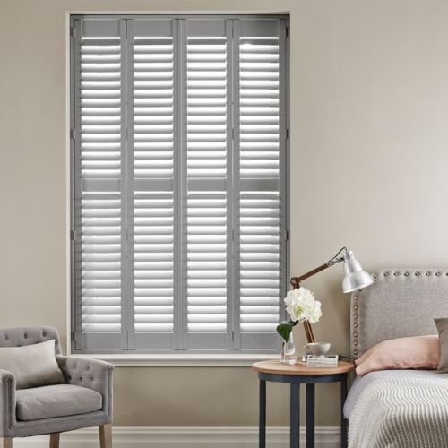 White plantation-style shutters covering a tall window, fully closed with slats slightly tilted to admit soft light; bedroom setting with upholstered bed, side table lamp, armchair, neutral walls.