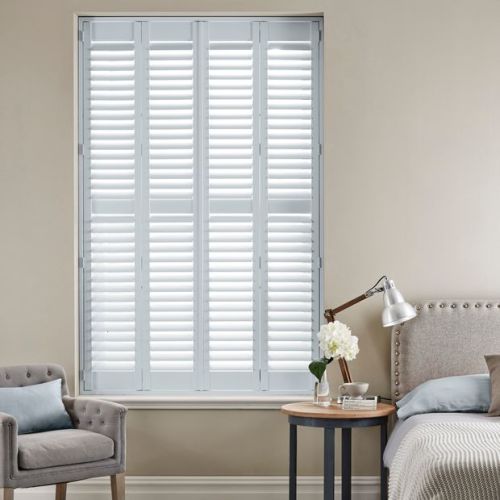 White full-height plantation shutters (four narrow panels) closed over the window, horizontal louvers slightly open and filtering bright daylight into a neutral-toned bedroom with armchair, bedside table, and lamp.