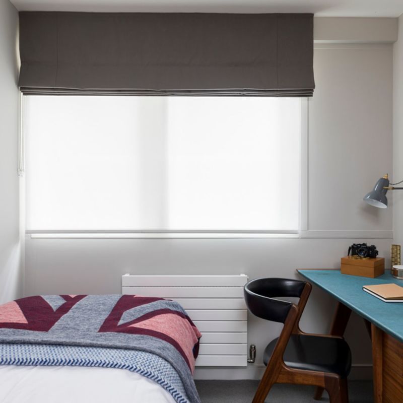 Grey Roman valance above a white roller blind; valance folded, roller fully lowered, filtering bright light into a small bedroom with a bed, desk and chair.
