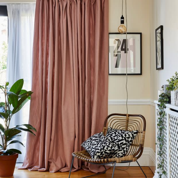 Dusty-rose floor-length curtains with sheer white voile behind, pleated and pooling; filtering daylight from a sliding door â€” corner living room with rattan chair, black-and-white cushions, plant, hanging bulb, framed 24 7