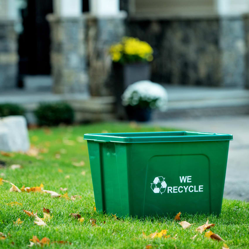green recycling container with we recycle stamped on the side sitting on a residential front lawn with leaves on the grass