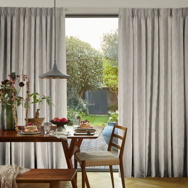 Floor-to-ceiling pleated beige curtains, partially drawn and filtering soft daylight through a central glass door; dining table with plates, pendant lamp and chairs, opening onto a verdant backyard.