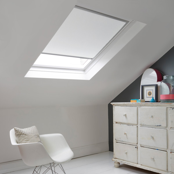 Room with white eaves wall, dark blue-grey wall to right with a shabby chic white drawer unit, a contemporary white chair, white painted floorboards and a skylight blind in white.