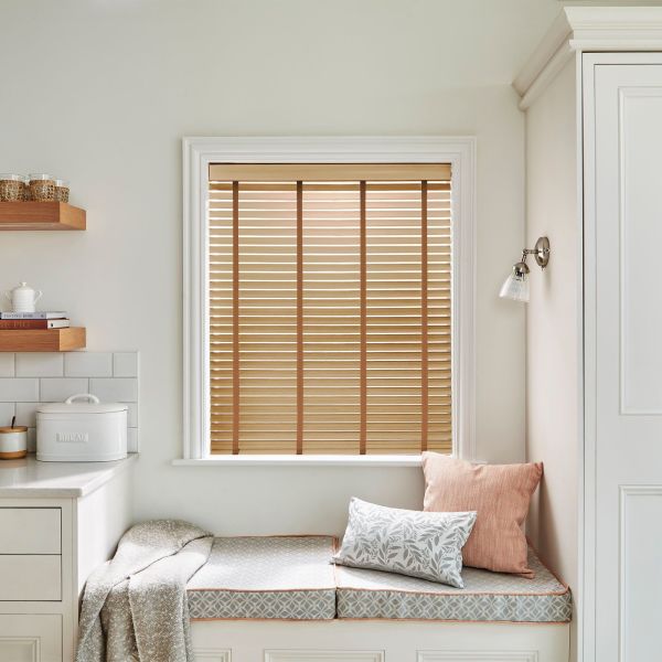 Wooden horizontal blinds with vertical tapes across the face, fully lowered and mostly closed, filtering warm daylight in a pale kitchen window-seat nook with cushions.
BREAD