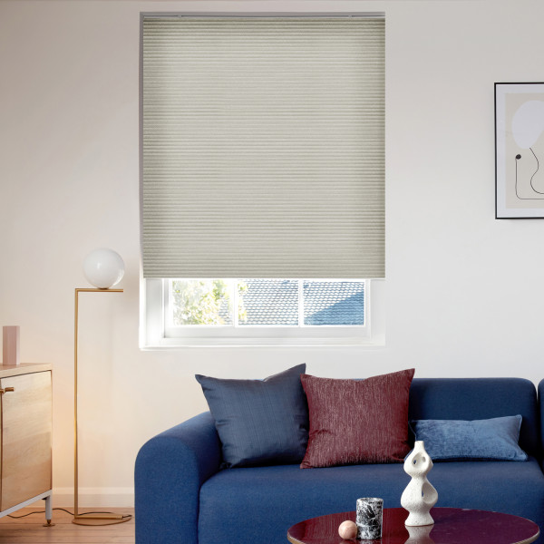 A beige cellular blind, lowered with horizontal ribs, filtering daylight over a sash window showing a roof and trees, in a living room with a blue sofa and lamp.