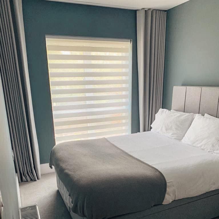 Layered zebra roller blind fills window, flanked by floor-length grey curtains; blind partially filters soft daylight while curtains are drawn open. Small bedroom with bed, teal walls and bedding.