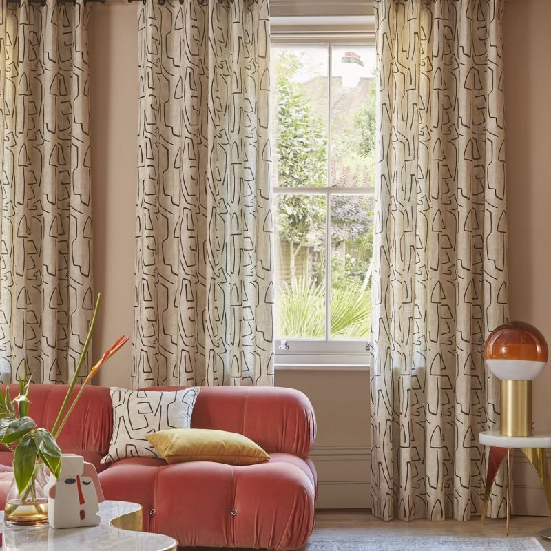 Floor-length patterned linen curtains, hung on a rod and drawn partly aside, filtering daylight and framing a sash window; living room with coral tufted sofa, side table and garden view.