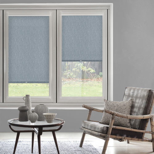 Blue textured roller blinds covering twin windowsâ€”left fully lowered, right partially loweredâ€”filtering light; living room with grey walls, wooden armchair, patterned cushion, side table and garden view.