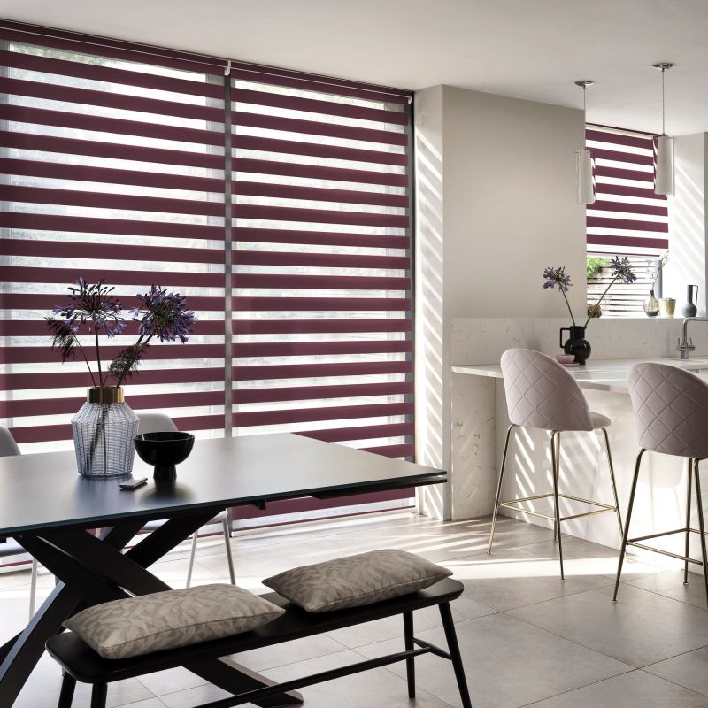 Dual-layer roller blinds with alternating burgundy opaque and sheer horizontal stripes covering floor-to-ceiling windows and a smaller kitchen window; filtering light into striped shadows; modern open-plan kitchen-dining.