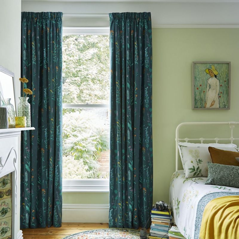 Teal patterned pleated curtains, hung and mostly closed at a tall sash window, filtering soft daylight into a pale-green bedroom with floral bedding, framed painting, wooden floor, and garden view.