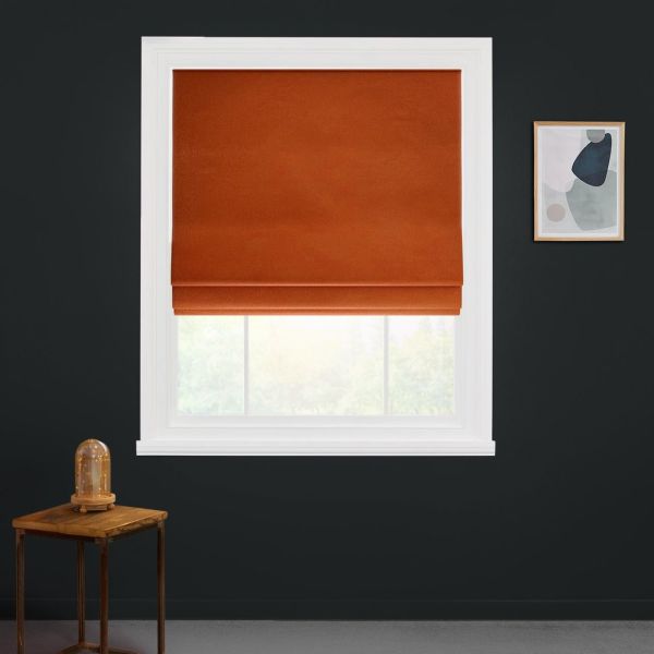 Rust-orange Roman blind with two folded tiers, lowered to cover the upper window, filtering daylight into a minimalist room — white-trimmed window set in a dark charcoal wall, small wooden side table.