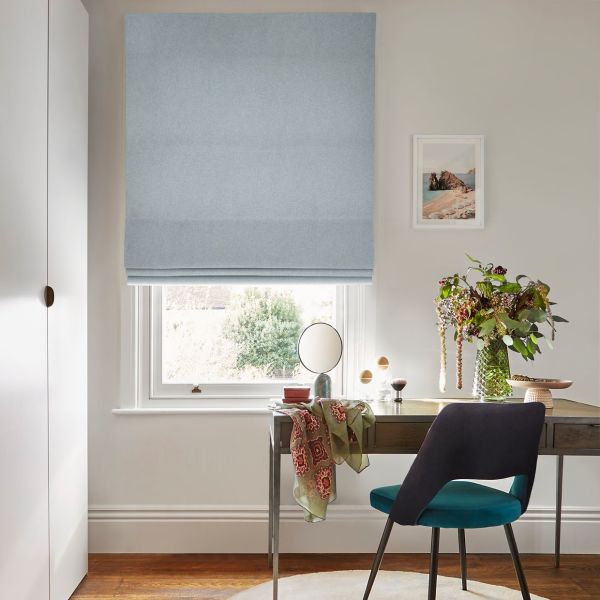 Pale blue Roman blind, lowered in soft folds, filtering daylight through a narrow bottom gap; desk beneath with mirror, vase of flowers and a teal-upholstered chair in a neutral study.