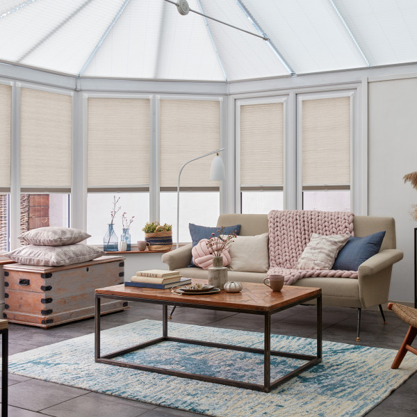 Beige pleated cellular blinds pulled down over tall conservatory windows, softly filtering daylight; cozy sunlit living room with sofa draped in a chunky pink knit, wooden coffee table, blue rug.