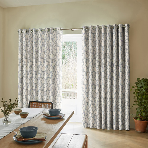 Floor-length grommet-top curtains with pale geometric blue-gray pattern, hung across a sliding glass door, partly drawn to reveal a narrow bright outdoor view; light-filled dining room with table and potted plant.