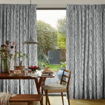 Gray textured floor-length curtains, hung to either side of a glass door, partially drawn and framing a sunlit garden; dining table set for breakfast, pendant lamp and wooden chairs inside.