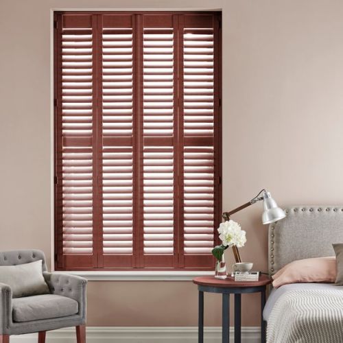 Wooden plantation shutters cover a tall recessed windowâ€”four-panel, mahogany louversâ€”mostly closed with slats angled to admit thin daylight; bedroom setting with upholstered bed, bedside table, lamp and armchair.