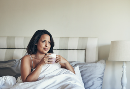 A woman sitting up in bed under a white blanket with a mug in her hands.