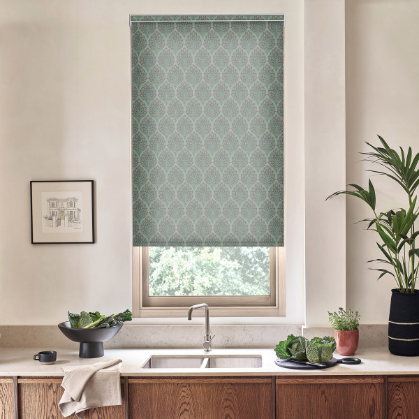 Patterned teal roller blind covers window above stainless sink, filtering light; kitchen countertop holds bowl of leafy greens, folded towel, potted plants, framed sketch on pale wall, wooden cabinets below.