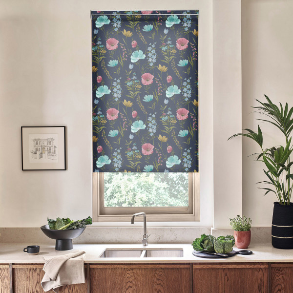Floral roller blind covers a window, displaying teal and pink blooms; light filters beneath. Stainless faucet and sink below, countertop with bowls of greens, framed sketch left, tall plant right.
