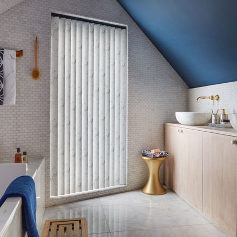 Vertical fabric blinds covering a window, closed and diffusing daylight; in bright bathroom with sloped blue ceiling, marble floor, bathtub left, vanity and vessel sink right.
