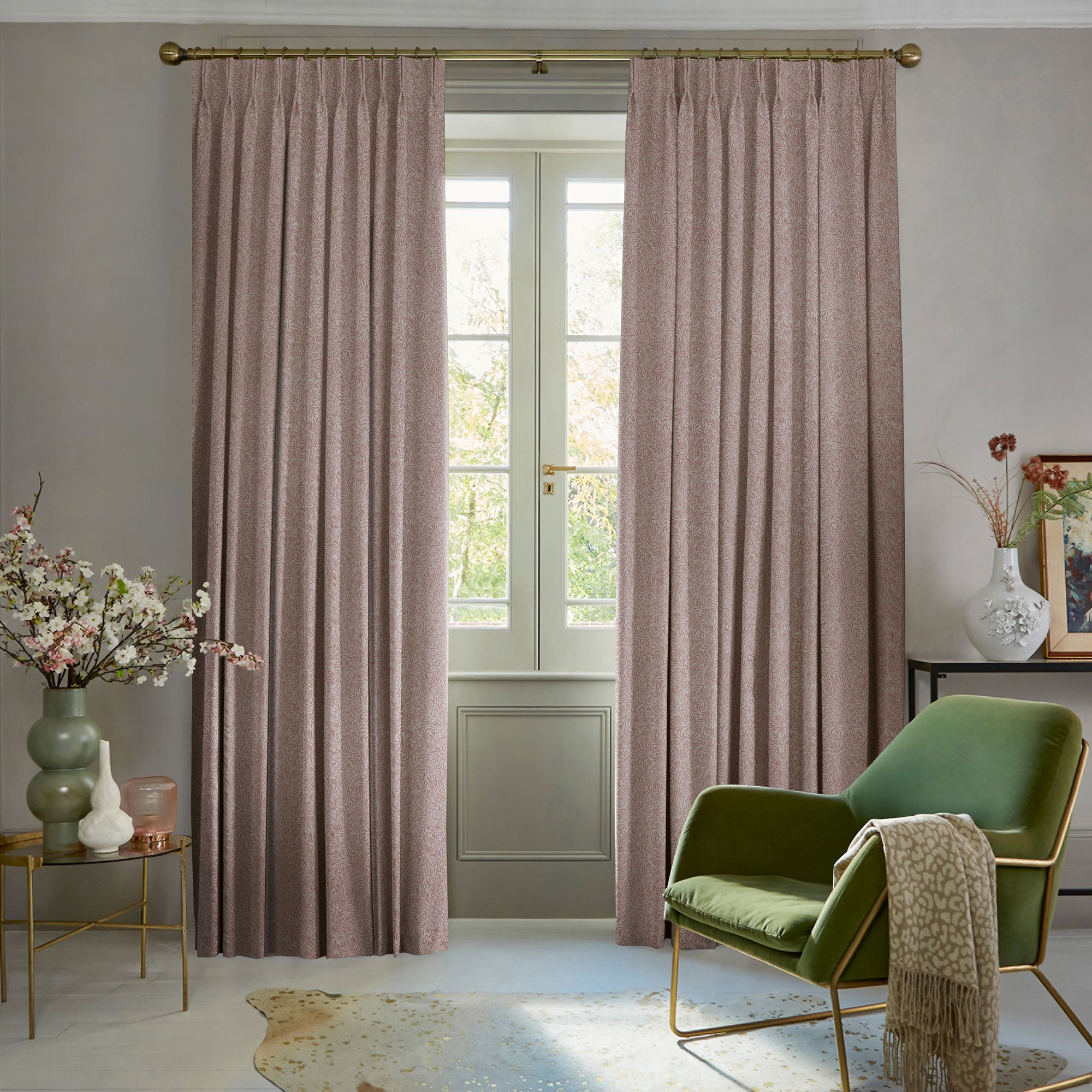 Pleated dusty-rose curtains on a brass rod, parted to reveal a glazed French window; softly filtering daylight in a serene living room with a green armchair and floral vases.