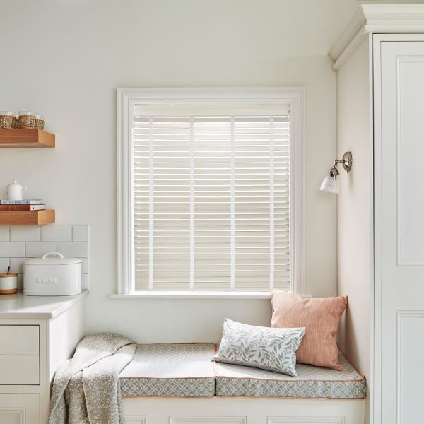 White horizontal venetian blinds covering the window, fully lowered with narrow slats filtering soft daylight; kitchen nook with built-in bench, patterned cushions, throw, wooden shelves, bread box, sconce.
BREAD