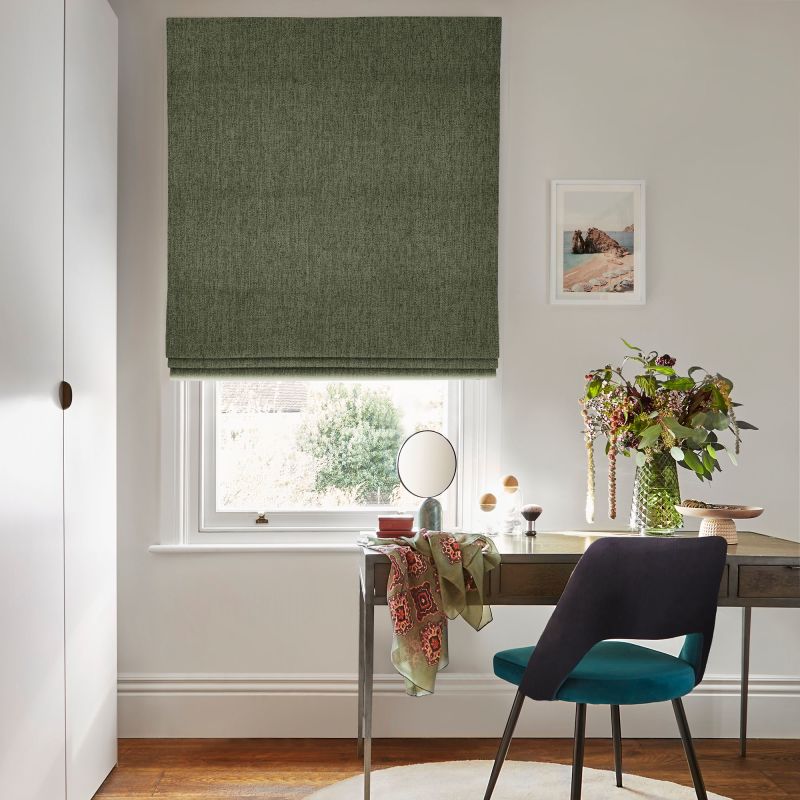 A green Roman blind, fully lowered with slight folds, covering a sash window and filtering soft daylight; desk beneath with vase of flowers and teal-upholstered chair in bright study.