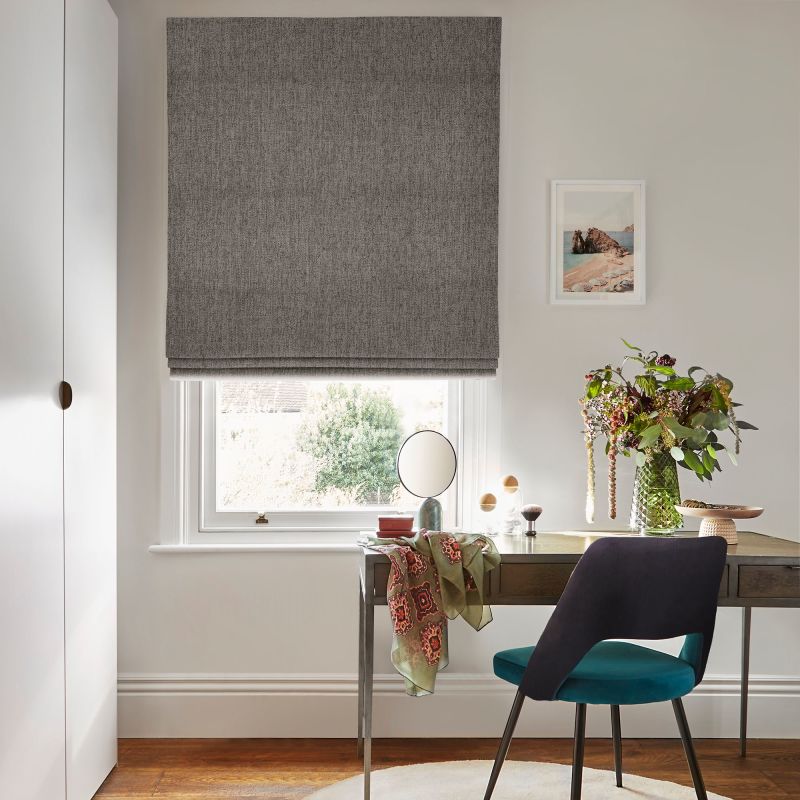 Grey fabric Roman blind lowered over a white sash window, filtering light; a wooden desk beneath holds a mirror, scarf, vase of flowers and teal-upholstered chair in a lit study.