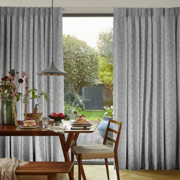 Floor-length grey pleated patterned curtains, parted at center to frame the view. A sunlit dining area with wooden table set for breakfast, pendant lamp, and a leafy backyard with shed.
