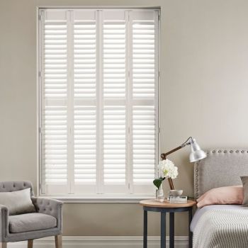 White plantation shutters covering a tall recessed window—four narrow hinged panels with horizontal louvers—closed, filtering soft daylight; neutral bedroom setting with upholstered armchair, bedside table, lamp and headboard.