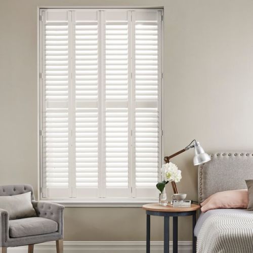 White plantation shutters covering a tall recessed windowâ€”four narrow hinged panels with horizontal louversâ€”closed, filtering soft daylight; neutral bedroom setting with upholstered armchair, bedside table, lamp and headboard.