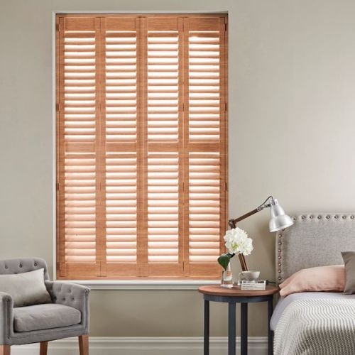 Wooden plantation shutters covering a tall window, closed with angled slats filtering warm daylight; set in a neutral bedroom corner with upholstered bed, bedside table, reading lamp and armchair.